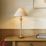Aldmont Avalon Table Lamp with clear glass column, antique brass base and cap, and tapered natural linen shade, shown on a wooden sideboard.