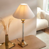 Aldmont Avalon table lamp with clear glass column, antique brass base and cap, and tapered linen shade, lit on a wooden side table.