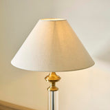 Aldmont Avalon Table Lamp with clear glass column, antique brass base and cap, and a tapered natural linen shade, shown against a light background.