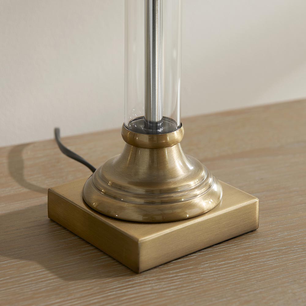 Close-up of the Aldmont Avalon Table Lamp base, showing a clear glass column set into an antique brass fitting on a square brass foot, on a wooden surface.