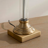 Close-up of the Aldmont Avalon Table Lamp base, showing a clear glass column set into an antique brass fitting on a square brass foot, on a wooden surface.