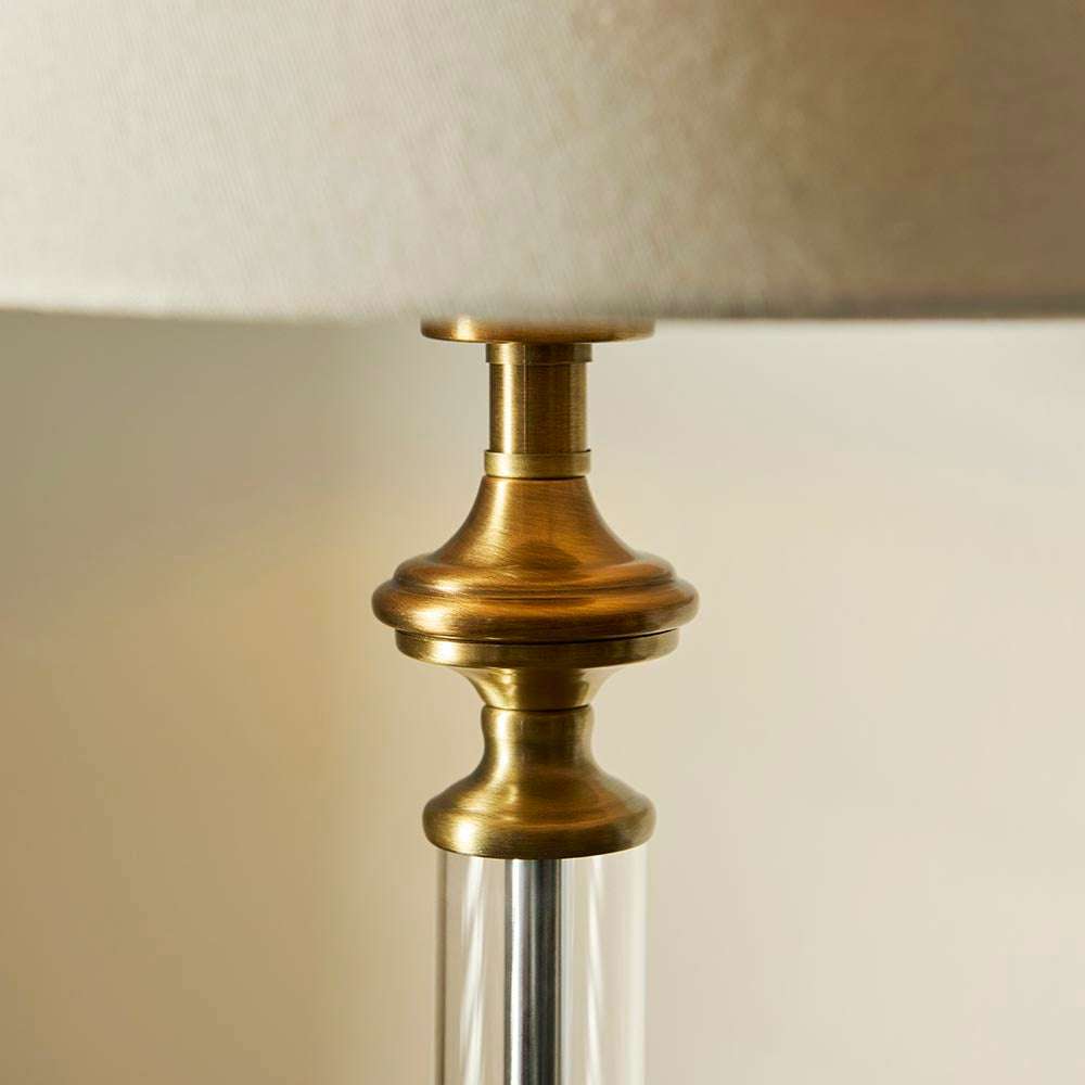 Aldmont Avalon Table Lamp close-up showing antique brass metalwork, clear glass column, and tapered natural linen shade edge, on a neutral background.
