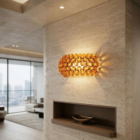 The Briller LED Large Wall Light in gold features amber glass elements, emitting warm LED light that creates decorative patterns on a light stone wall in a minimalist living room with large windows and city views.