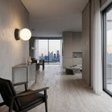 Minimalist apartment with light wood floors, neutral walls, modern black chair, Origo Concrete Wall Light in black & white, and large windows framing a city skyline. Simple décor gives the space an airy, uncluttered feel.