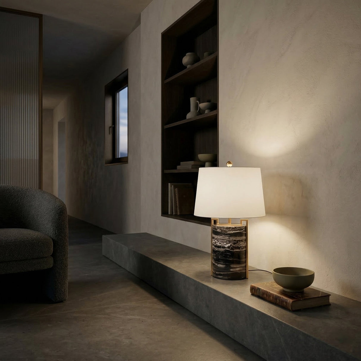 A modern minimalist interior features a stone bench with the Obrion 1 Light Framed Marble Table Lamp in Brass & Black, a bowl, and a book. Built-in shelves display ceramics, while soft lighting creates a cozy, serene mood.