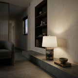 A modern minimalist interior features a stone bench with the Obrion 1 Light Framed Marble Table Lamp in Brass & Black, a bowl, and a book. Built-in shelves display ceramics, while soft lighting creates a cozy, serene mood.