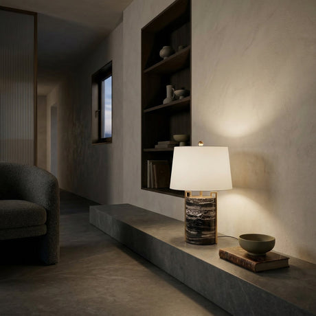 A modern minimalist interior features a stone bench with the Obrion 1 Light Framed Marble Table Lamp in Brass & Black, a bowl, and a book. Built-in shelves display ceramics, while soft lighting creates a cozy, serene mood.