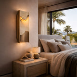A modern bedroom with a big window and palm trees, a neutral bed, a stone nightstand with a lamp and vases, and the Veloris LED Small Carved Alabaster Wall Light in Brushed Brass & Soft White adding warm, marble-like accent lighting.
