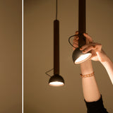 Berry S Aluminium LED pendant light with slim bronze tube and angled domed head, shown in hand and hanging against a warm neutral wall.