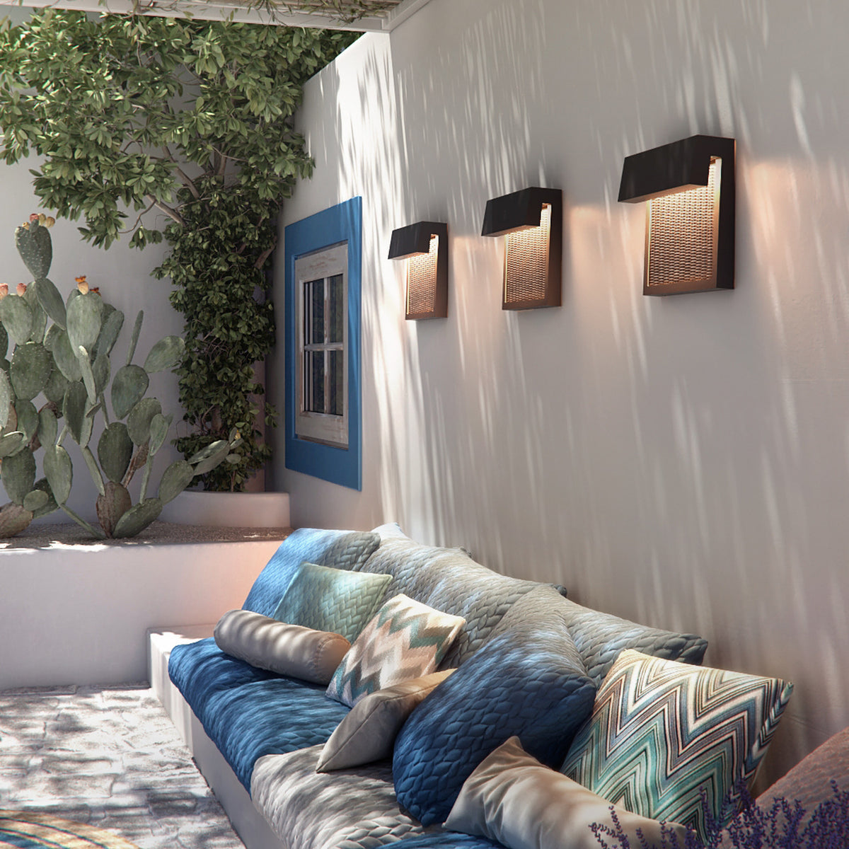 Sisal A/01 LED outdoor wall lights in brown, three fitted on a textured wall above a patio sofa with patterned cushions, each with a woven fibre front panel and angled hood casting downward light.