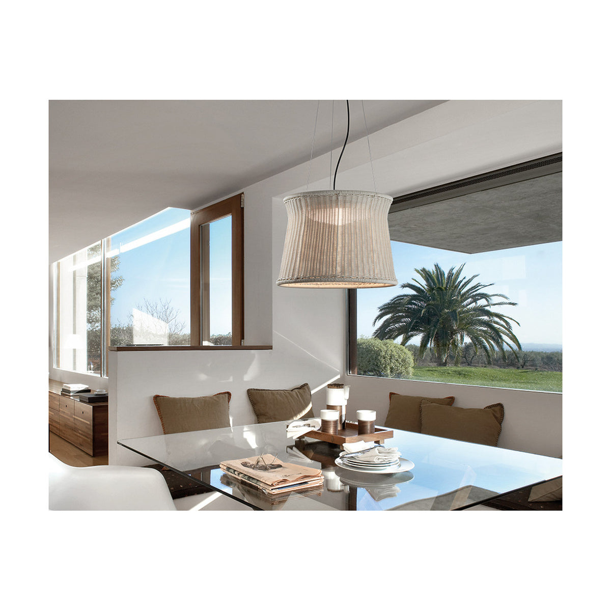 Syra 60 Aluminium LED Outdoor Pendant Light with ivory white woven drum shade, waisted shape, hung above a glass dining table in a sunlit room with garden views.