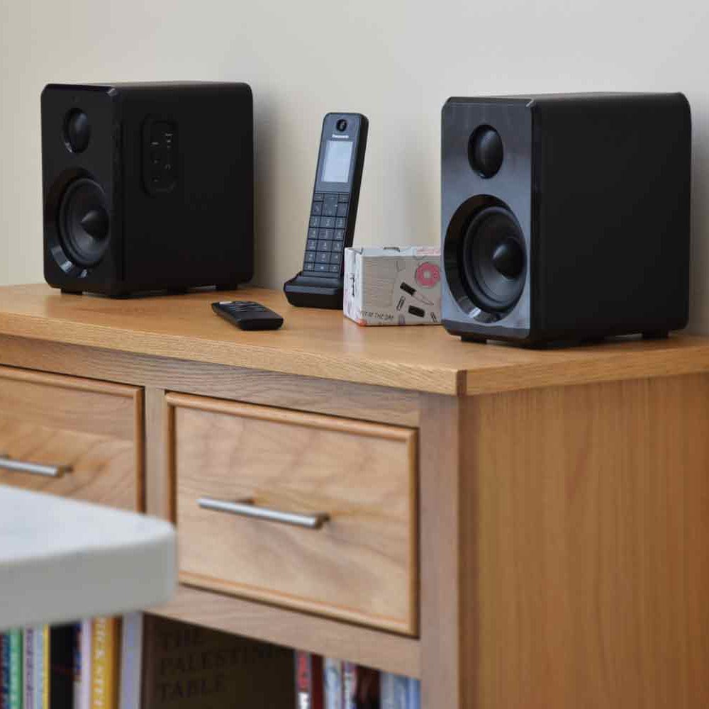 AVSL AV:Link ABS35BLK active Bluetooth bookshelf speakers in matt black on a wooden sideboard, shown with the included remote alongside a phone and small box.