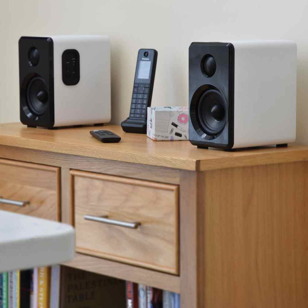 AV:Link ABS35WHT Active Bluetooth Bookshelf Speakers by AVSL in satin white with black driver panels, shown on a wooden sideboard beside a remote and phone.