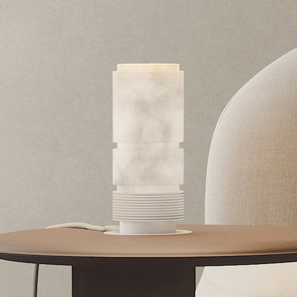 Bowie LED Cylinder Alabaster Table Lamp with stacked banded column and ribbed white cap on an iron base, softly lit on a side table beside a cream armchair.
