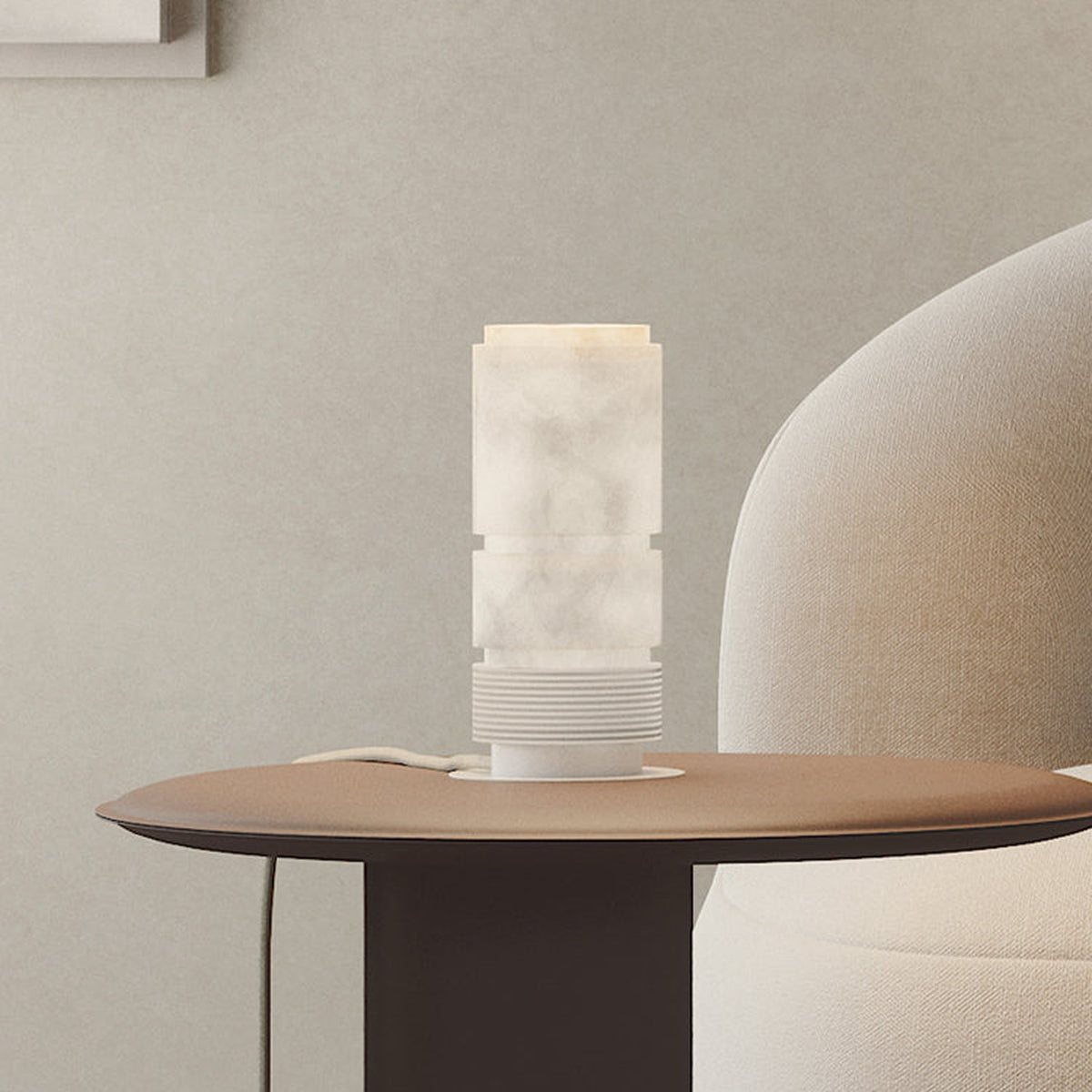 Bowie LED Cylinder Alabaster Table Lamp with stacked column shade, ribbed white cap, and iron base glowing softly on a side table beside a cream armchair.