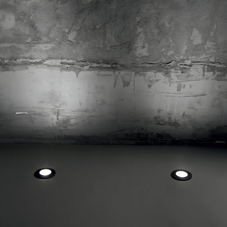 A dimly lit ceiling with an industrial design incorporates two circular lights, similar to the Brass Large Round Outdoor Recessed Ground Light in grey, near the base. Above, a textured, unfinished concrete surface is illuminated by dimmable lighting that accentuates the rough ceiling texture.