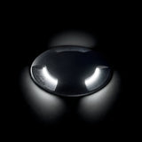 The Venus Outdoor Large Drive Over Recessed Light - Black is a dark circular fixture set amidst black space, showcasing two curved, bright white lights reminiscent of eyes or headlights. It has an IP67 rating and casts reflections on the surface below.