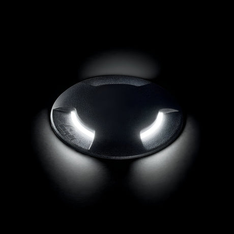 The Venus Outdoor Large Drive Over Recessed Light - Black is a dark circular fixture set amidst black space, showcasing two curved, bright white lights reminiscent of eyes or headlights. It has an IP67 rating and casts reflections on the surface below.