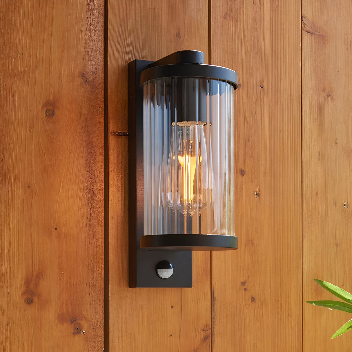 Endon Castbury matt black outdoor wall light with ribbed glass cylinder, E27 filament bulb and PIR sensor, mounted on a timber plank wall.