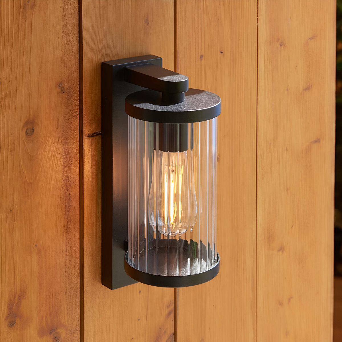 Endon Castbury outdoor wall light in matt black with a cylindrical clear ribbed glass shade and E27 filament bulb, mounted on a timber clad wall.