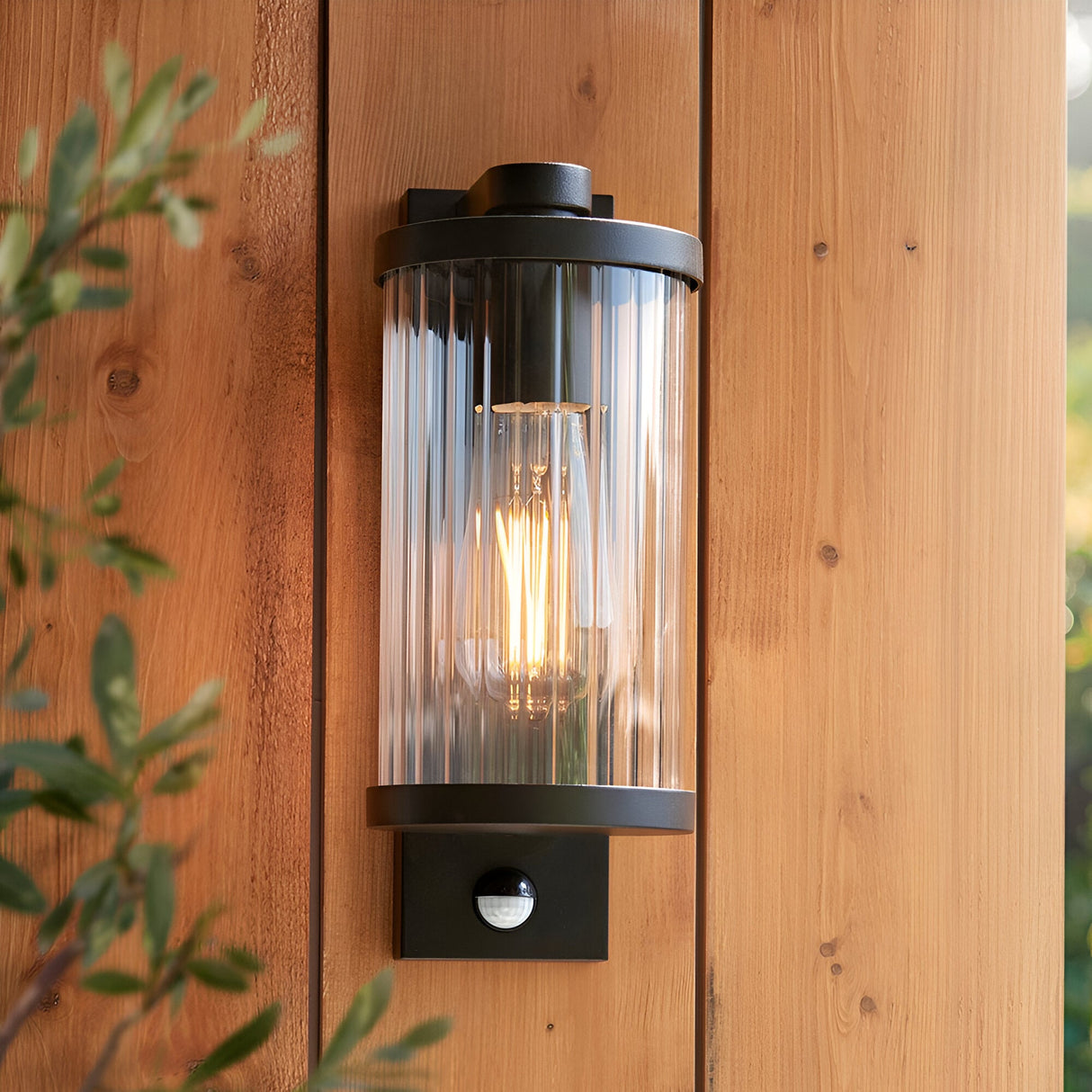 Endon Castbury matt black outdoor wall light with ribbed glass cylinder, E27 filament bulb and PIR sensor, mounted on a timber wall beside foliage.