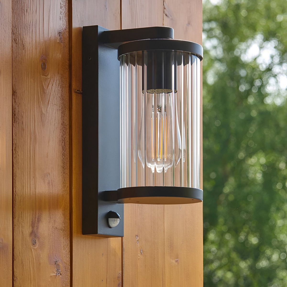 Endon Castbury matt black outdoor wall light with ribbed clear glass shade, E27 filament bulb and PIR sensor, mounted on a timber clad wall.