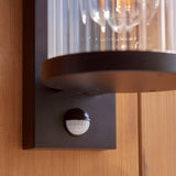 Close-up of Endon Castbury outdoor wall light in matt black, showing the PIR sensor beneath the ribbed glass cylinder on a timber wall.