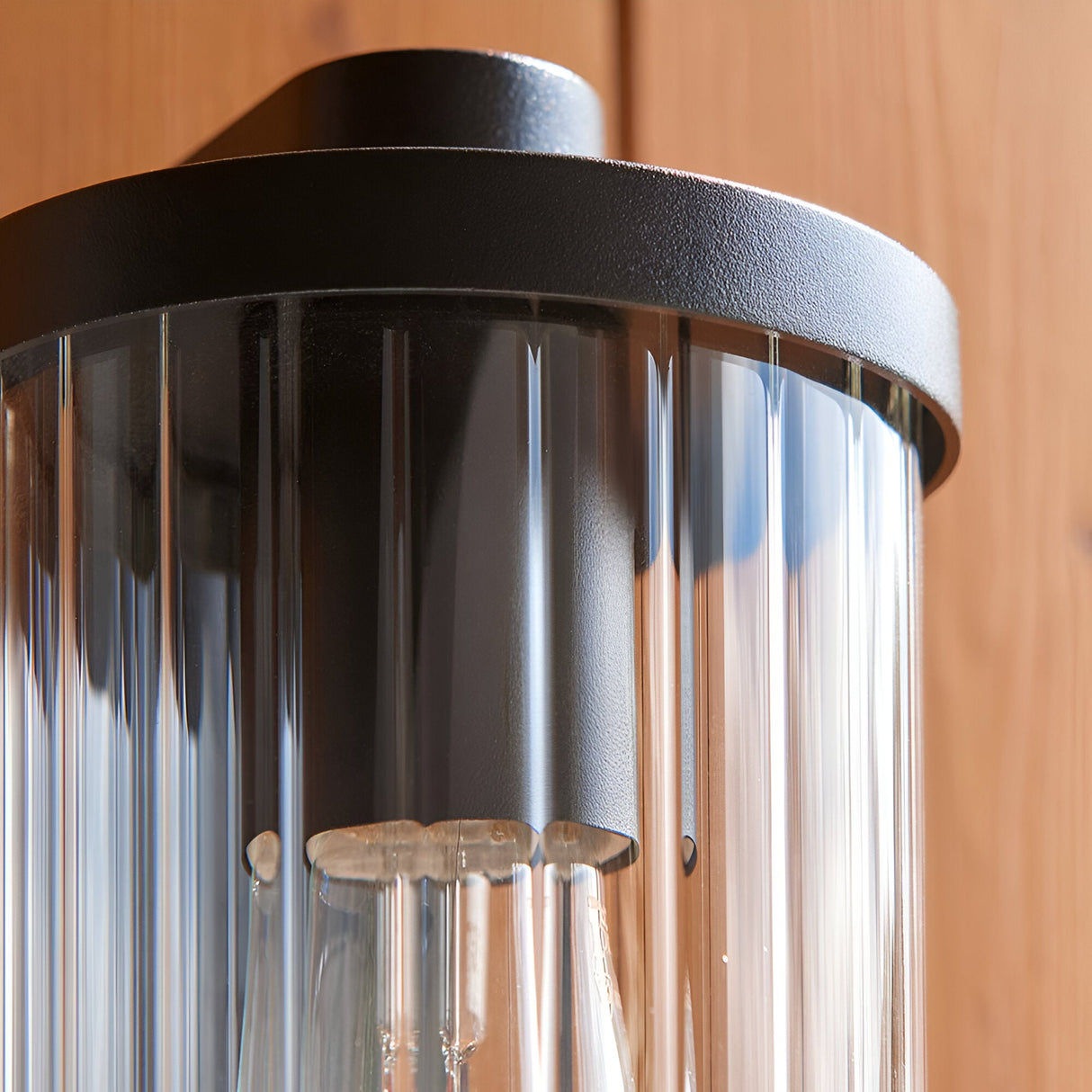 Close-up of Endon Castbury outdoor wall light showing the matt black cylindrical top and clear ribbed glass shade mounted on a timber wall.