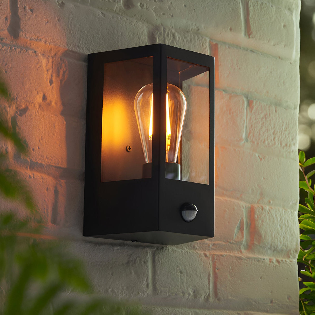 Brenshaw Outdoor Wall Light in matt black, cuboid frame with clear glass panels, PIR motion sensor, and E27 bulb, mounted on a brick wall.