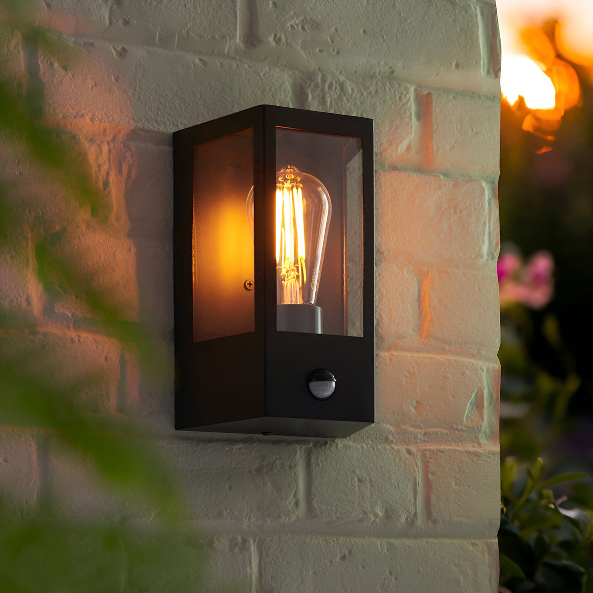 Brenshaw Outdoor Wall Light in matt black with a cubed metal frame, clear glass panels, PIR motion sensor and E27 bulb, mounted on a brick wall at dusk.