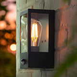 Brenshaw Outdoor Wall Light in matt black, cubed steel frame with clear glass panels, PIR sensor, glowing E27 bulb, mounted on a brick wall at dusk.