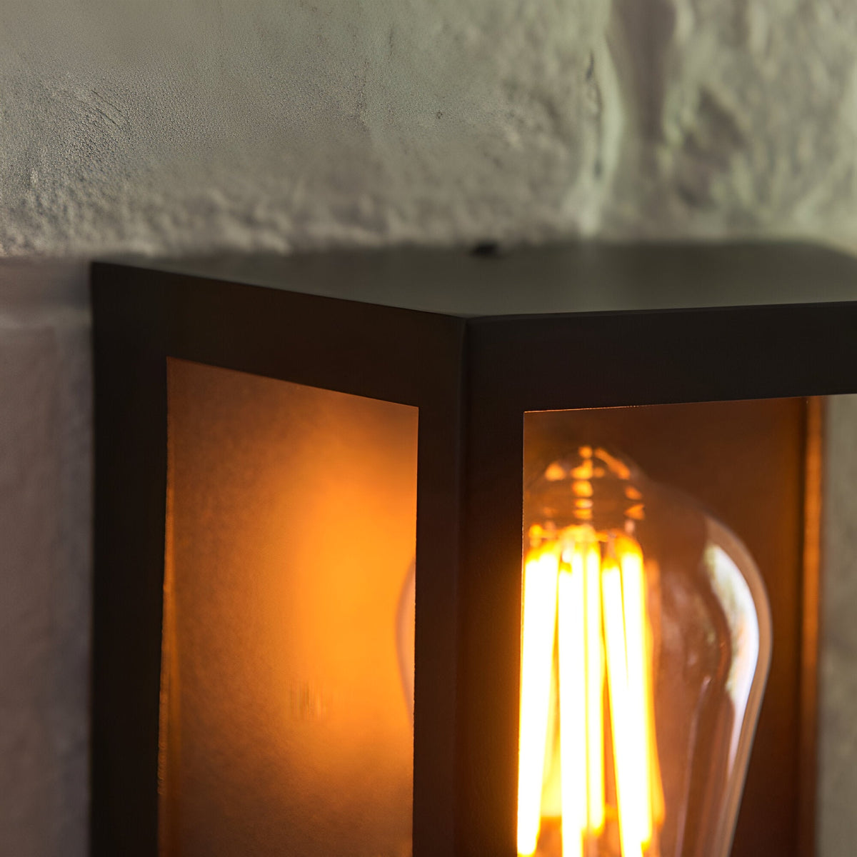 Close-up of the Brenshaw Outdoor Wall Light in matt black, showing its cubed metal frame, clear glass panels, and lit E27 bulb against a textured wall.