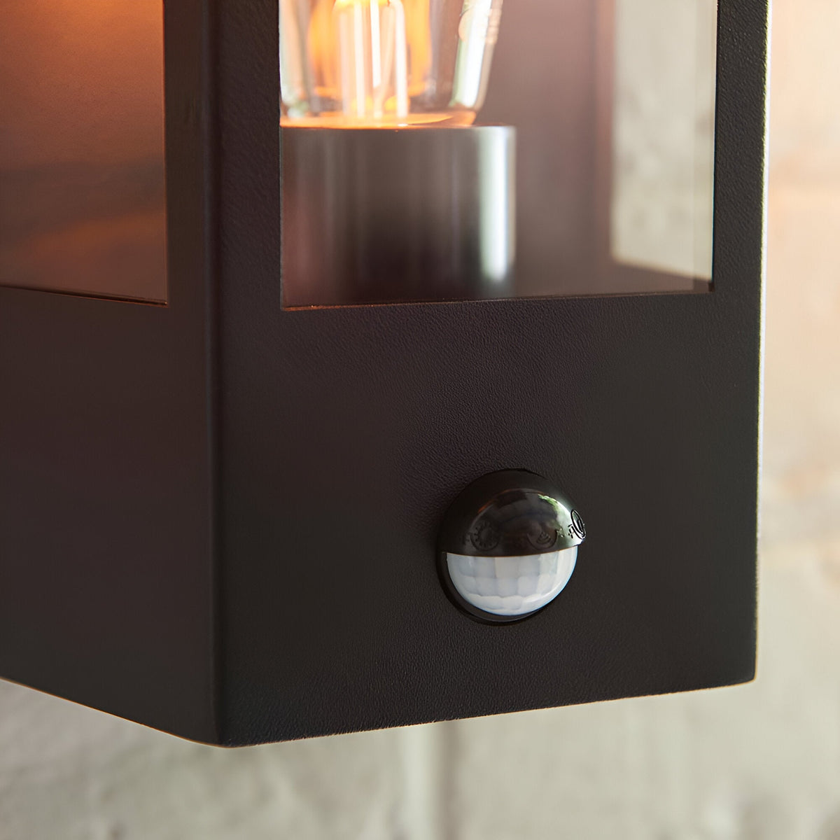 Close-up of the Brenshaw Outdoor Wall Light in matt black with clear glass panel, visible bulb, and integrated round PIR motion sensor on the base.