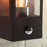 Close-up of the Brenshaw Outdoor Wall Light in matt black with clear glass panel, visible bulb, and integrated round PIR motion sensor on the base.