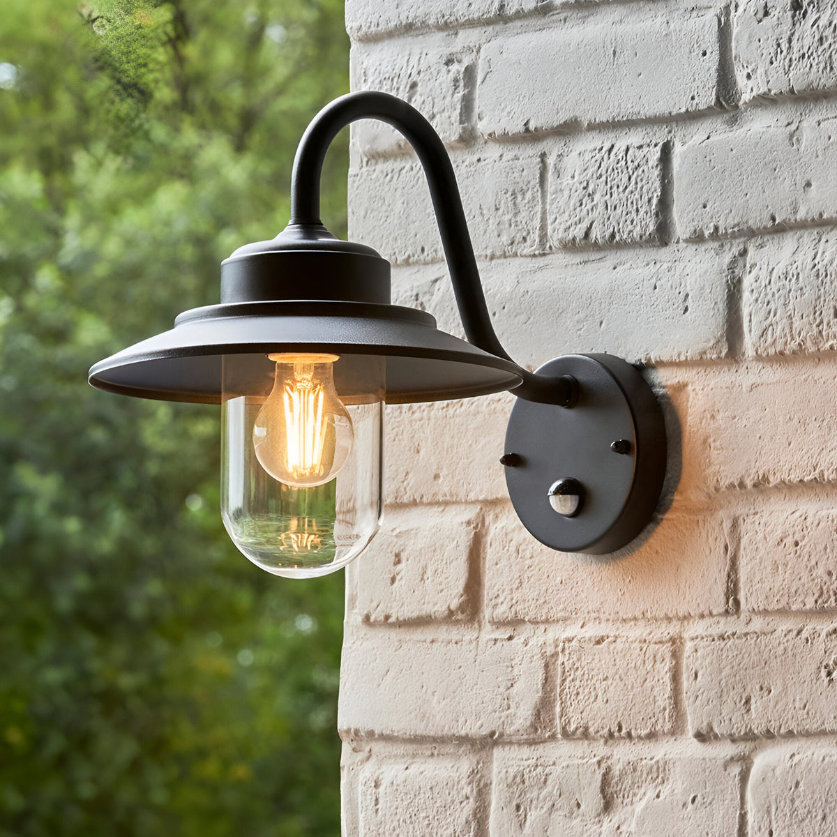 Endon Cranhurst matt black outdoor wall light with curved arm, wide-brimmed shade, clear glass dome, E27 filament bulb and PIR sensor on a white brick wall.