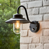 Endon Cranhurst matt black outdoor wall light with curved arm, wide-brimmed shade, clear glass dome, E27 filament bulb and PIR sensor on a white brick wall.