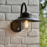 Endon Cranhurst matt black outdoor wall light with curved arm, wide-brim shade, clear glass dome and PIR sensor, mounted on a white brick wall.