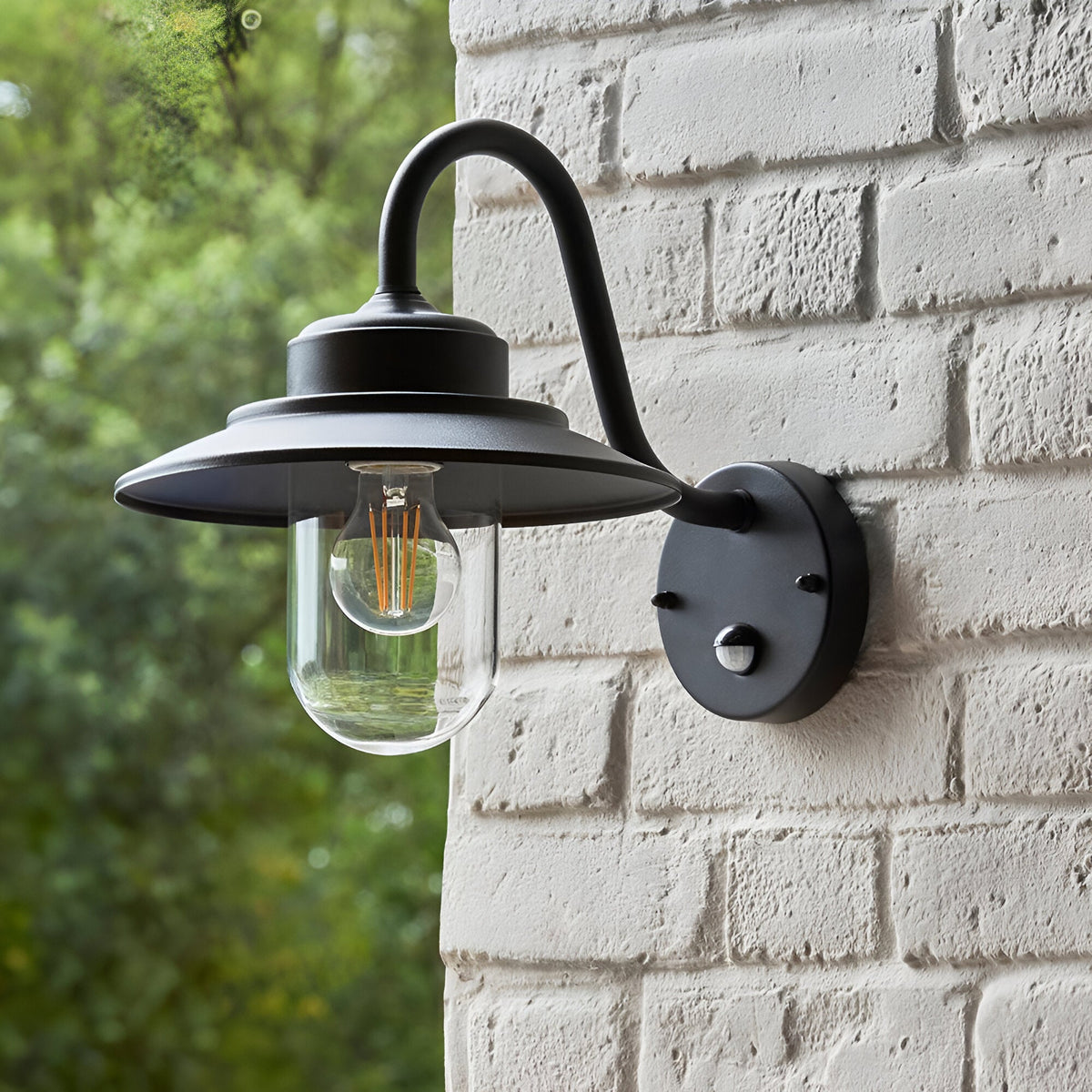Endon Cranhurst outdoor wall light in matt black with curved arm, wide-brimmed shade, clear glass dome, E27 filament bulb and PIR sensor on a brick wall.