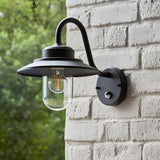 Endon Cranhurst outdoor wall light in matt black with curved arm, wide-brimmed shade, clear glass dome, E27 filament bulb and PIR sensor on a brick wall.