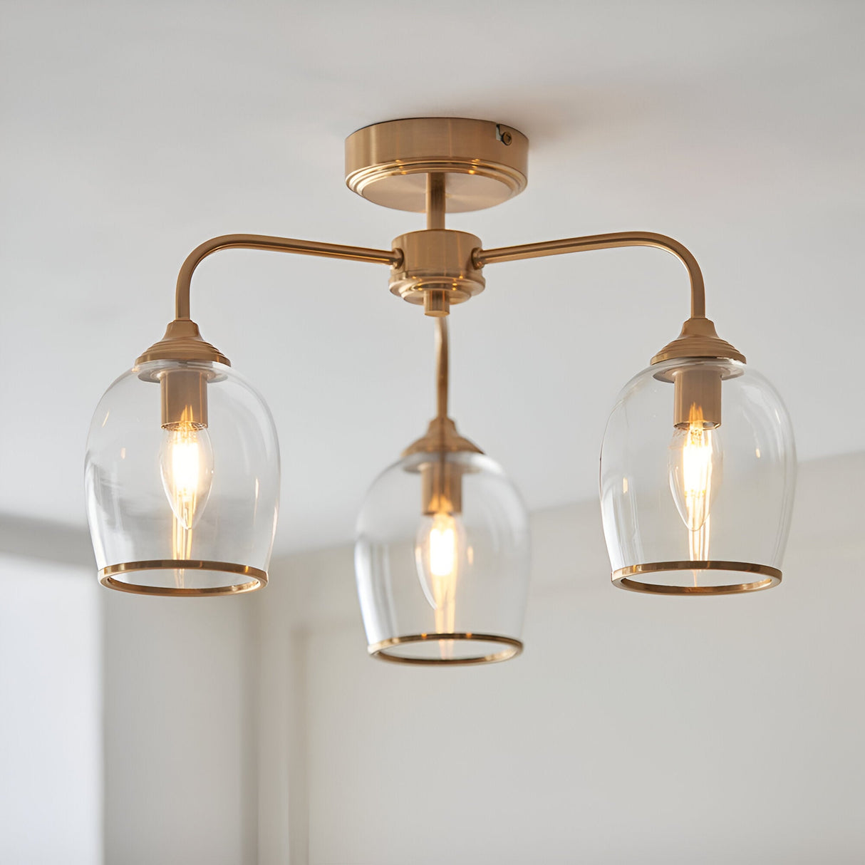 Halstead 3-light semi-flush ceiling light in aged brass, with curved arms holding clear glass shades and exposed bulbs, mounted on a white ceiling.
