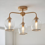 Halstead 3-light semi-flush ceiling light in aged brass, with curved arms holding clear glass shades and exposed bulbs, mounted on a white ceiling.