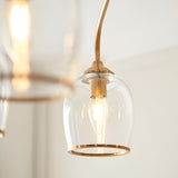 Halstead 5 Light Pendant in brass, close-up of a clear glass shade with brass trim, arched brass arm, and filament bulb, other shades softly blurred behind.