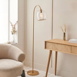 Halstead Floor Lamp in aged brass with curved stem and clear domed glass shade, shown beside an armchair and wooden desk in a living room setting.