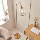 Halstead Floor Lamp in aged brass with curved stem and domed clear glass shade, shown beside a wooden side table in a living room setting.