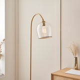 Halstead Floor Lamp in aged brass with a curved stem and domed clear glass shade, shown in a living room beside a wooden cabinet.