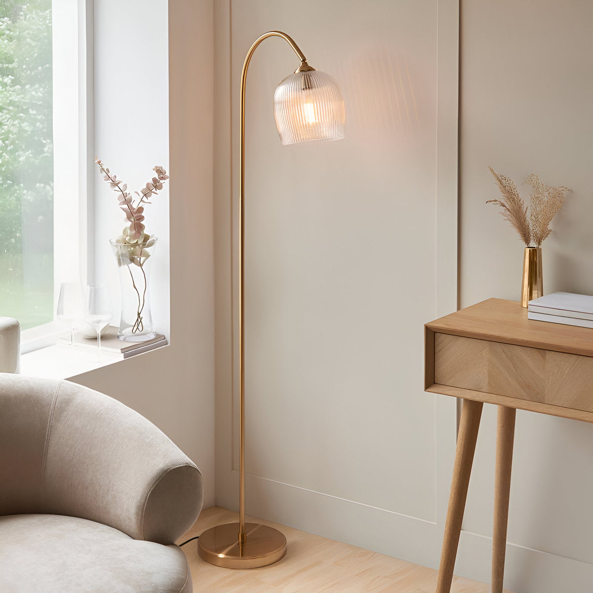 Endon Halstead floor lamp in aged brass with a curved stem and ribbed clear glass domed shade, beside a cream armchair and oak console table.