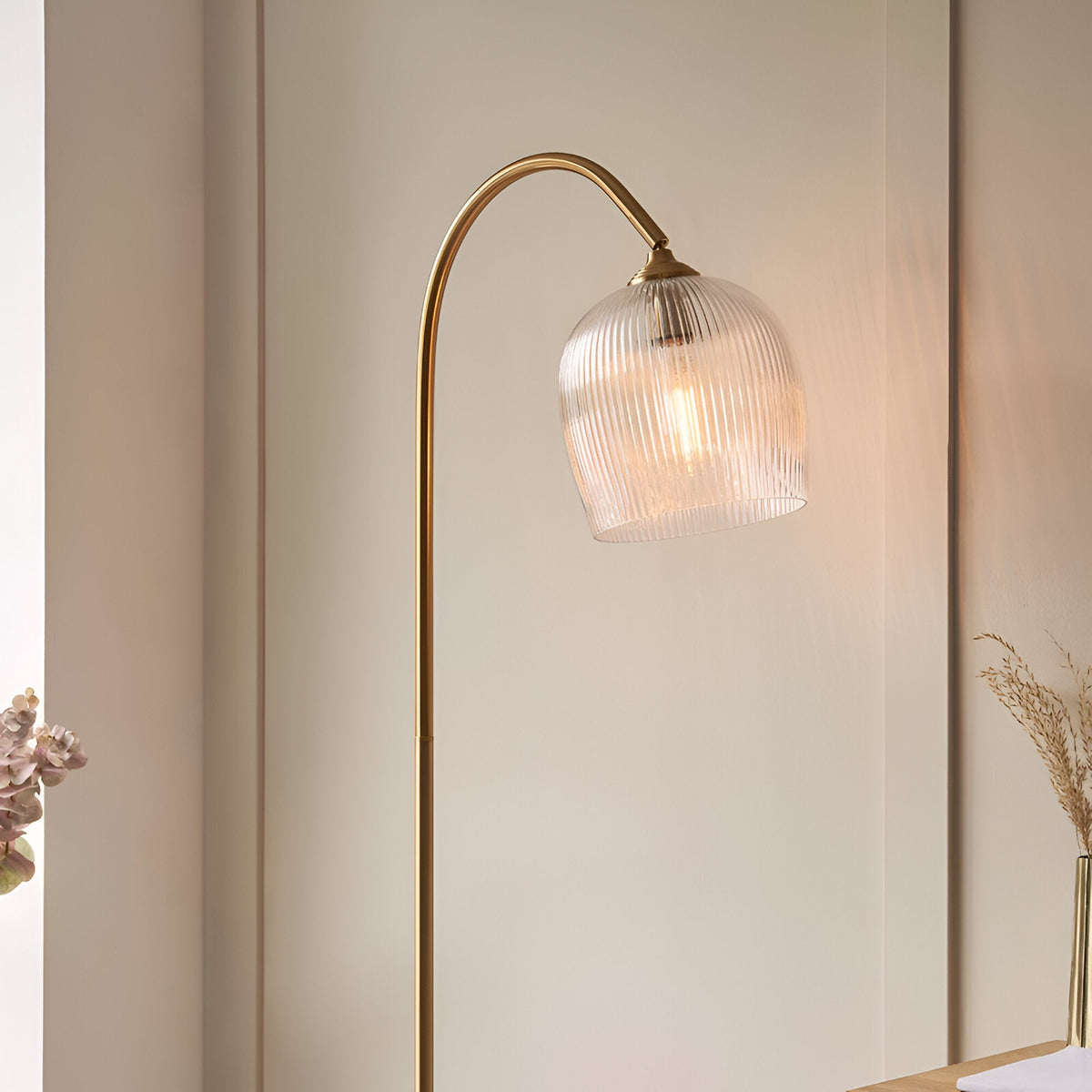 Endon Halstead floor lamp with arched aged brass stem and ribbed clear glass domed shade, lit beside a neutral panelled wall and dried floral stems.