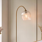 Endon Halstead floor lamp with arched aged brass stem and ribbed clear glass domed shade, lit beside a neutral panelled wall and dried floral stems.