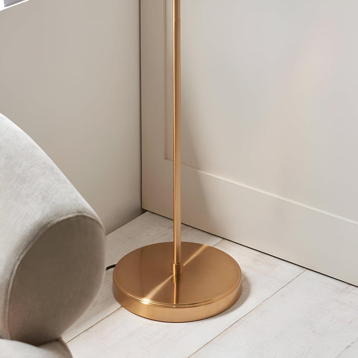Close-up of the Endon Halstead floor lamp's round aged brass base and brushed stem on pale wood flooring beside a neutral upholstered chair.