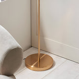 Close-up of the Endon Halstead floor lamp's round aged brass base and brushed stem on pale wood flooring beside a neutral upholstered chair.